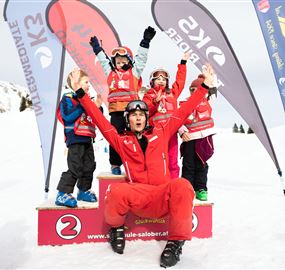 Ein fröhlicher Skilehrer steht auf dem Podium und feiert mit Kindern, die Medaillen tragen. Im Hintergrund sind Schneelandschaften und Flaggen zu sehen.