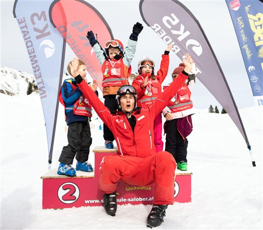 A group of children in ski gear stands on a podium in the snow. A ski instructor joyfully poses with them while flags wave in the background.