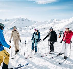 Eine Gruppe von Skifahrern steht auf einer schneebedeckten Piste in den Alpen. Der Himmel ist klar und die Berge sind im Hintergrund sichtbar.