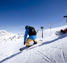 Zwei Snowboarder fahren über die schneebedeckte Piste. Im Hintergrund sind Berge und ein klarer blauer Himmel zu sehen.