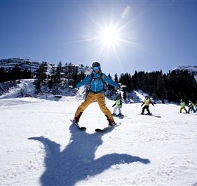 A sunny winter landscape with slopes and a group of skiers. In the foreground, there is a snowboarder in a blue jacket and yellow pants.