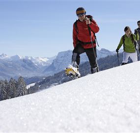 A group of three people is hiking through a snowy landscape. Mountains and a blue sky are visible in the background.