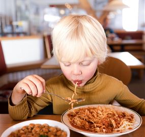 Ein Junge mit blonden Haaren isst Spaghetti und schaut konzentriert auf sein Essen. Neben ihm steht eine Schüssel mit Kichererbsen.
