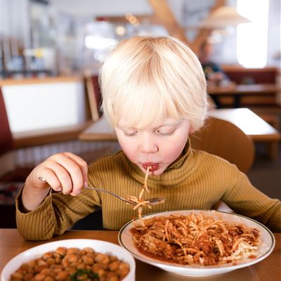 Ein Junge mit blonden Haaren isst Spaghetti und schaut konzentriert auf sein Essen. Neben ihm steht eine Schüssel mit Kichererbsen.