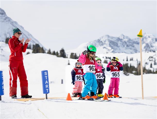 Eine Gruppe von Kindern in bunten Skianzügen steht an der Startlinie eines Skirennens. Ein Trainer applaudiere und motiviert die kleinen Skifahrer.