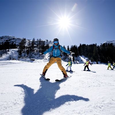 Ein Skifahrer steht auf einer schneebedeckten Piste unter einem klaren, blauen Himmel. Im Hintergrund sind weitere Skifahrer und Berge zu sehen.