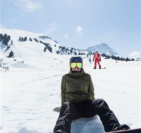 A snowboarder sits in the snow and enjoys the wintry mountain scenery. In the background, additional skiers are visible.