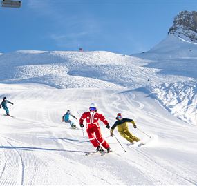 Eine Gruppe von Skifahrern fährt den schneebedeckten Hang hinunter. Der klare blaue Himmel und die Berge im Hintergrund schaffen eine perfekte Winterlandschaft.
