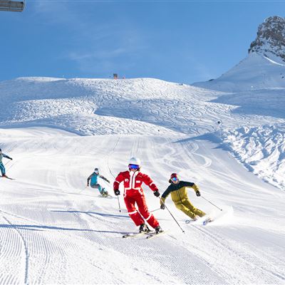 Skifahrer fahren auf einer verschneiten Piste unter blauem Himmel. Die Berge im Hintergrund bieten eine malerische Kulisse.