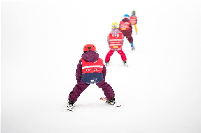 Eine Gruppe von Kindern in Skikleidung fährt im Schnee. Sie tragen rote Westen mit der Aufschrift "BORN TO SKI".