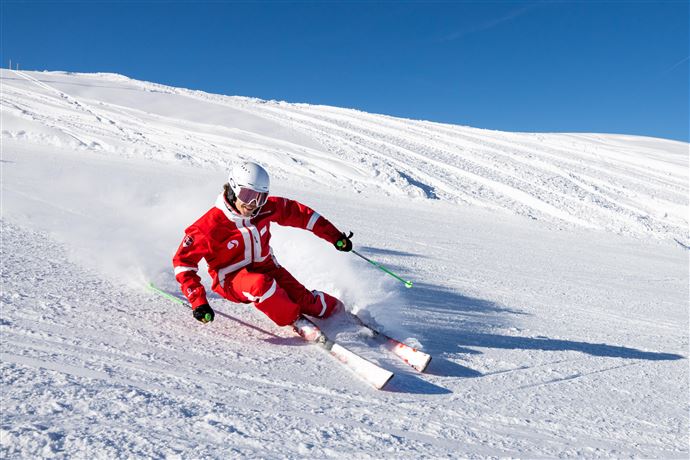 Ein Skifahrer in roter Kleidung fährt mit Schwung über die verschneite Piste. Der Himmel ist klar und blau.