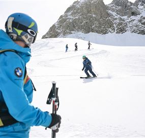 Eine Gruppe von Skifahrern genießt den frischen Schnee in den Bergen. Im Vordergrund steht ein Skilehrer in blauer Ausrüstung.