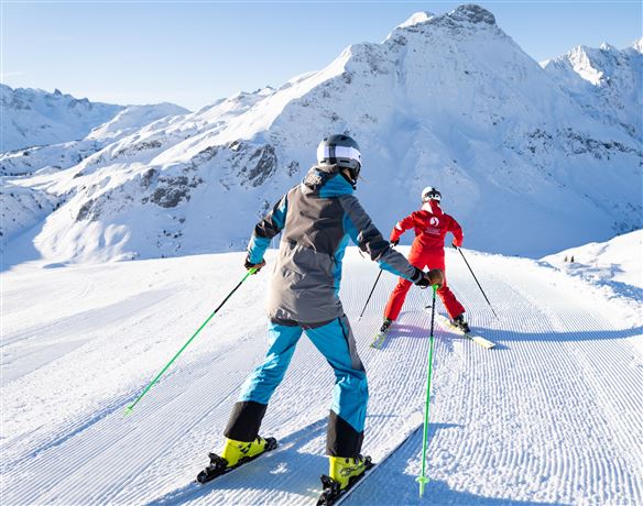 Zwei Skifahrer auf einer schneebedeckten Piste in den Bergen. Die Sonne scheint und die Landschaft ist beeindruckend.