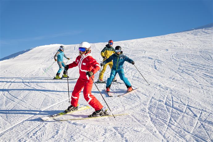 Vier Skifahrer bei sonnigem Wetter auf einer verschneiten Piste. Sie tragen farbige Skiausrüstungen und genießen die Abfahrt.