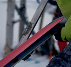 Ein Skifahrer hält ein Skisportgerät in der Hand. Im Hintergrund sind Schneelandschaften zu sehen.