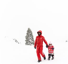 Eine Person im roten Anzug geht mit einem kleinen Kind durch den Schnee. Im Hintergrund ist ein einzelner, schneebedeckter Baum zu sehen.