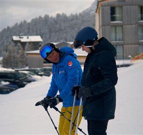 Two men are standing in the snow and speaking to each other. In the background, there are snow-covered mountains and buildings visible.