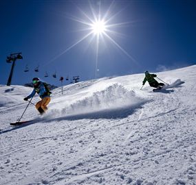 Two skiers are gliding down a snow-covered slope beneath a bright blue sky. The sun is shining brightly over the winter landscape.