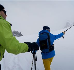 Zwei Personen in warmer Kleidung wandern durch eine schneebedeckte Landschaft. Der eine hält einen Stock, während sie Richtung Berg schauen.