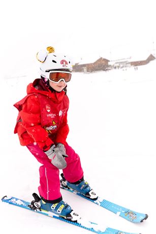 Ein Kind fährt Ski in einem verschneiten Gebiet. Es trägt eine rote Jacke, eine Skibrille und einen Helm mit Ohren.