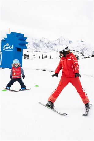 Eine Skilehrerin in roter Kleidung zeigt einem kleinen Schüler das Skifahren. Beide stehen auf einem schneebedeckten Hang mit Bergen im Hintergrund.