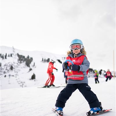 Ein jungen Skifahrer auf einer schneebedeckten Piste, der voller Freude posiert. Im Hintergrund sind andere Skifahrer und eine Berglandschaft zu sehen.