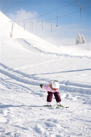 Ein Kind lernt das Skifahren auf einer schneebedeckten Piste. Im Hintergrund sind Skilifte und ein blauer Himmel zu sehen.