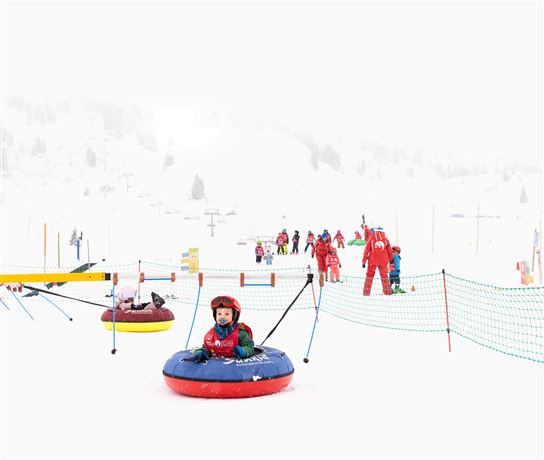 Ein Winterspaß im Schnee mit Kindern, die in Reifen rutschen. Im Hintergrund sind Skilehrer und weitere Kinder zu sehen.