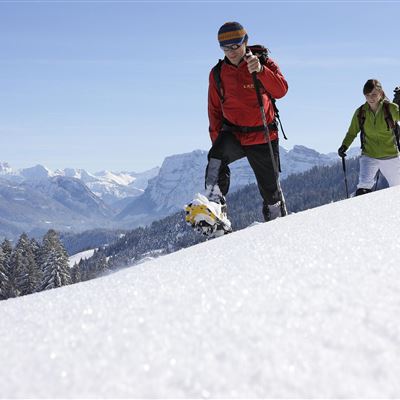 Drei Personen wandern durch den Schnee in den Bergen. Die Sonne scheint und der Himmel ist klar.