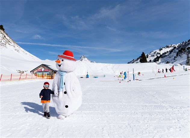 Ein Schneemann mit einem rot-weißen Hut steht neben einem Kind in einem verschneiten Skigebiet. Im Hintergrund sind weitere Kinder und Spielgeräte zu sehen.