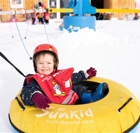 Ein fröhliches Kind sitzt in einem gelben Reifen im Schnee. Im Hintergrund sind andere Kinder und eine schneebedeckte Landschaft zu sehen.
