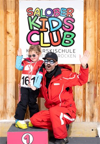 Ein fröhliches Kind und ein Skilehrer posieren lächelnd vor einem bunten Schild des „Salober Kids Club“. Beide heben die Hände und tragen Sonnenbrillen, während sie auf einem Podest stehen.