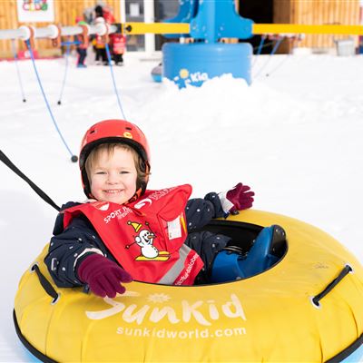 Ein fröhliches Kind sitzt in einem gelben Reifen im Schnee. Im Hintergrund sind andere Kinder und eine schneebedeckte Landschaft zu sehen.