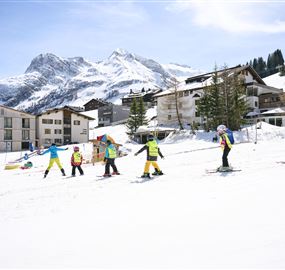 Eine Gruppe von Skifahrern in bunten Outfits fährt über einen schneebedeckten Hang. Im Hintergrund sind Berge und eine kleine Ansiedlung zu sehen.