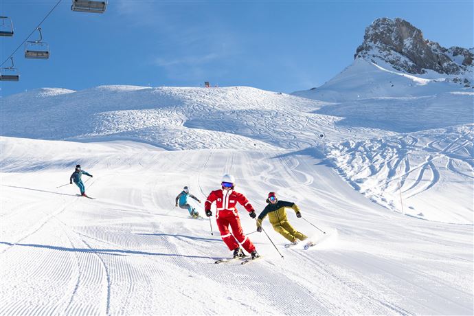 Skifahrer genießen eine sonnige Abfahrt auf einer schneebedeckten Piste. Im Hintergrund sind majestätische Berge zu sehen.