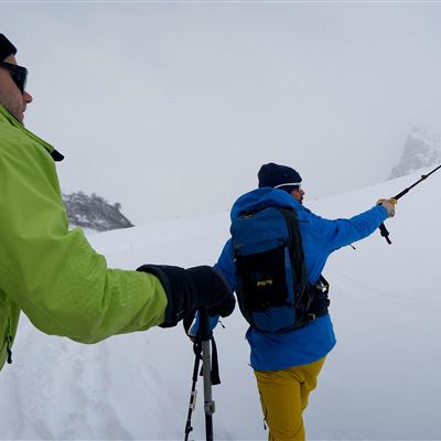 Zwei Personen wandern durch verschneite Landschaften. Der eine trägt eine grüne Jacke und der andere eine blaue Jacke und verwendet einen Stock.