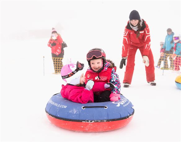 Zwei Kinder sitzen fröhlich in einem Schlitten im Schnee. Eine Aufsichtsperson steht in der Nähe und schaut zu.