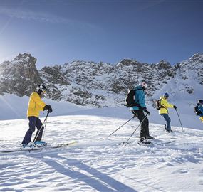 Eine Gruppe von Skifahrern fährt durch eine schneebedeckte Landschaft. Im Hintergrund sind beeindruckende Berge und ein strahlender Himmel zu sehen.