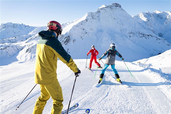 Drei Skifahrer genießen das sonnige Wetter auf einer frisch präparierten Piste in den Bergen. Im Hintergrund sind schneebedeckte Gipfel zu sehen.