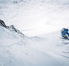 A skier jumps over a snow-covered slope in the mountains. The sky is cloudy, and the landscape appears wintry and impressive.
