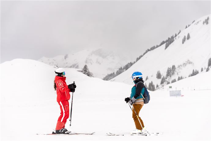 Zwei Skifahrer stehen auf einer verschneiten Piste. Im Hintergrund sind schneebedeckte Berge unter einem grauen Himmel zu sehen.