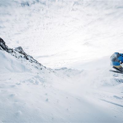 A skier jumps over a snow-covered slope in the mountains. The sky is cloudy, and the landscape appears wintry and impressive.