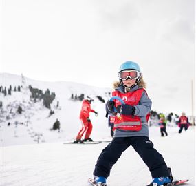 A small child is skiing on a snow-covered slope. In the background, there are other skiers and a snowy landscape visible.