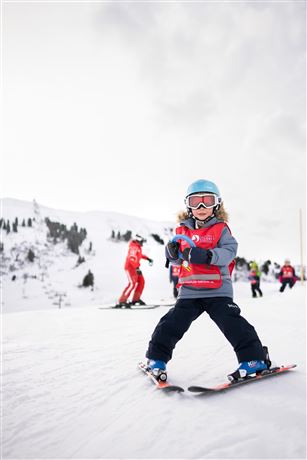 A small child is skiing on a snow-covered slope. In the background, there are other skiers and a snowy landscape visible.