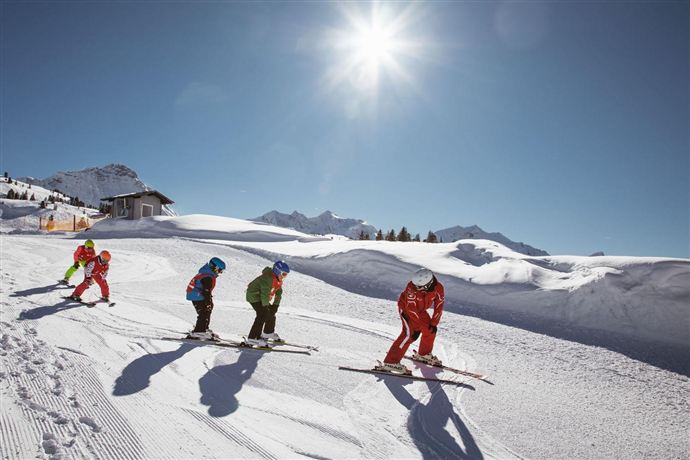 A group of skiers is skiing on a closed slope in the sun. In the background, snow-covered mountains and a building are visible.