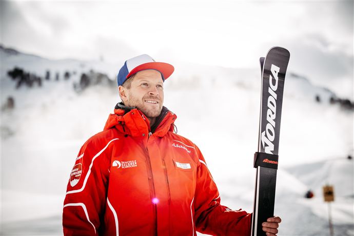 Ein Skilehrer steht in einem winterlichen Bergsetting. Er trägt eine rote Jacke und hält ein Paar Skier in der Hand.