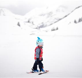Ein kleines Kind fährt Ski in einer schneebedeckten Landschaft. Die Berge im Hintergrund sind ebenfalls mit Schnee bedeckt und der Himmel ist bewölkt.