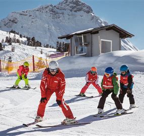 Een groep skiërs leert op een besneeuwd skipiste. Op de achtergrond zijn de bergen en een skischool te zien.