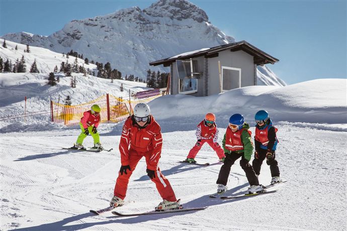 A group of skiers is learning on a snowy slope. In the background, the mountains and a ski school can be seen.