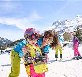Eine fröhliche Mutter umarmt ihr Kind auf einer Skipiste. Im Hintergrund sind schneebedeckte Berge und andere Skifahrer zu sehen.